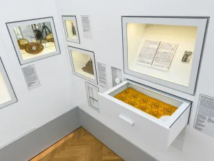 Ausstellungsraum: verschiedene Vitrinen mit historischen Artefakten, einem offenen Buch und einem gelben Tuch mit Davidsternen enthält