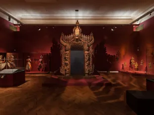 Ausstellungsraum: Geschnitzte hölzerne Türrahmen auf roter Plattform, rote Wände, Vitrinen mit Kostümen und Statuen