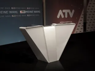 moderner Pulttisch im Fernsehstudio mit dem ATV-Logo und "Meine Wahl"-Schildern im Hintergrund