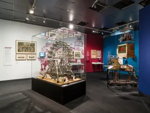 Ausstellungsraum: Modell eines Riesenrads in Vitrine, historische Bilder an roter und weißer Wand, Schaukelpferd