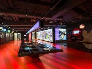Besucherzentrum mit roter Bodenfläche, einem langen Tisch mit interaktiven Displays und bunten Wänden mit Ausstellungsstücken über den Wiener Nahverkehr