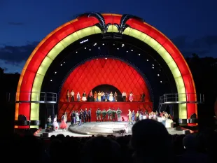 Szene: Bunte Freilichtbühne bei Nacht mit rot-gelber Rundung. Schauspieler in bunter Kleidung und Uniformen tanzen oder stehen auf einer Plattform, Publikum vor der Bühne