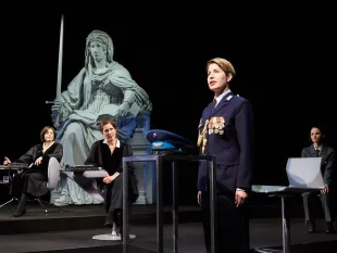 Szene: Eine Bühne mit einer Statue einer sitzenden Justitia mit Schwert und Waage, umgeben von Personen in Uniform und Roben, darunter eine mit Medaillen geschmückte Figur