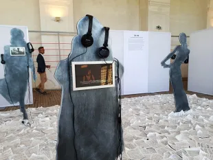 Ausstellungsraum mit abstrakten Silhouettenfiguren, die Kopfhörer tragen und kleine Bildschirme mit Fotos oder Videos enthalten.