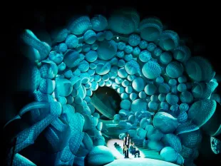 Szene: Drei Personen in einem Tunnel aus blauen, kugelförmigen Strukturen, die einen himmlischen Wolkenstrudel darstellen