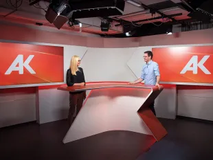 Ausstellungsraum: Modernes Studio mit rot-weißem Pult und "AK"-Logo an den Wänden. Zwei Personen stehen am Pult, beleuchtet von Deckenstrahlern