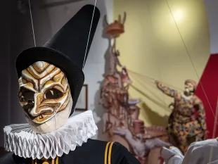 Ausstellungsraum: Marionette mit farbiger Maske, schwarzem Hut und weißem Kragen steht im Vordergrund. Im Hintergrund sind weitere bunte Marionetten