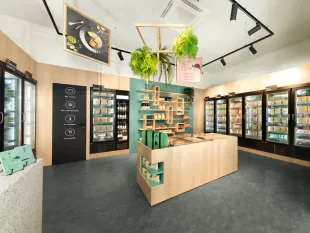 Supermarkt mit modernem Design, beleuchteten Kühlregalen mit gefrorenen Lebensmitteln, einer zentralen Holztheke mit Regalen und hängenden Pflanzen sowie einem Bild mit Speisen an der Decke.