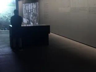 Person steht in einem schwach beleuchteten Ausstellungsraum, betrachtet eine Projektion und Paneele über die Distanz und Vernichtung der Deportierten.
