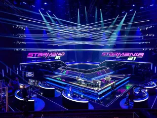 Moderne Bühne in blauem Licht, durchzogen von weißen Laserstrahlen, die sich kreuzen. Die mehrstufige Fläche mit leuchtenden Rändern trägt das "Starmania 21" Logo in Pink und Grün. Techniker bedienen runde Pultstationen mit Monitoren