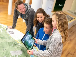 Ausstellungsraum: Famlie steht vor einem Landschaftsmodell. Kind hält ein Tablet mit Bild einer Landschaft