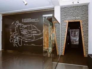 Ausstellungsraum mit einer großen Wandkarte, einer Vitrine mit Textil und einem „Glück Auf“-Eingang zu einem dunklen Stollen.