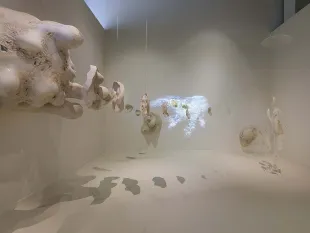 Überlebensgroße Amöbenskulptur und transparente menschliche Fragmente hängen in einem Raum, mit projizierten Mikroorganismen im Hintergrund.