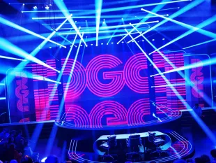 Leuchtende TV-Bühne mit großer LED-Wand im Hintergrund, die den Schriftzug „GGC“ zeigt, davor eine runde Plattform mit einem Tisch und drei Stühlen, durchzogen von Lichtstrahlen.