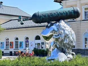 glänzende Skulptur einer Frau ab den Schultern, mit einer Sauerstoffflasche mit der Aufschrift "Atemluft" zentriert auf ihrem Kopf platziert, vor einem Gebäude mit Hecken und Blumen, gekennzeichnet durch ein Schild "Atemluft 2024 Xenia Hausner". Die Skulptur hat eine Höhe von etwa drei Metern und besteht aus poliertem Aluminium und Bronze.