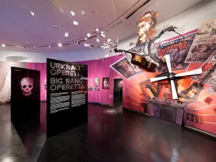 Ausstellungsraum: Pinker Raum mit Kontrabass-Skulptur, Mann, Plakaten wie "Moulin Rouge" und "Urknall Operetta", hängende Papierfetzen und Schädel