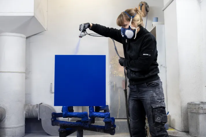 Frau mit Atemschutzmaske, die mit einer Lackierpistole einen blauen Würfel lackiert