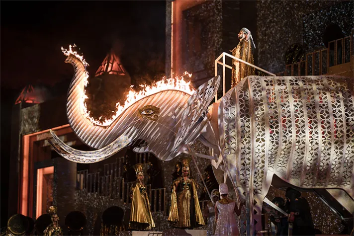 Bühnenbild von AIDA mit einem metallenen Elefantenmodell, dessen Rüssel in Flammen steht, begleitet von Figuren in goldenen Gewändern und einer detaillierten Kulisse.
