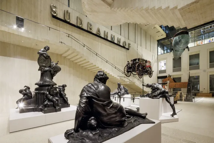 Innenansicht des Wien Museums mit monumentalen historischen Skulpturen im Vordergrund, einer hängenden Postkutsche und einem Walmodell im Hintergrund. An der Wand großer Schriftzug „Südbahnhof“
