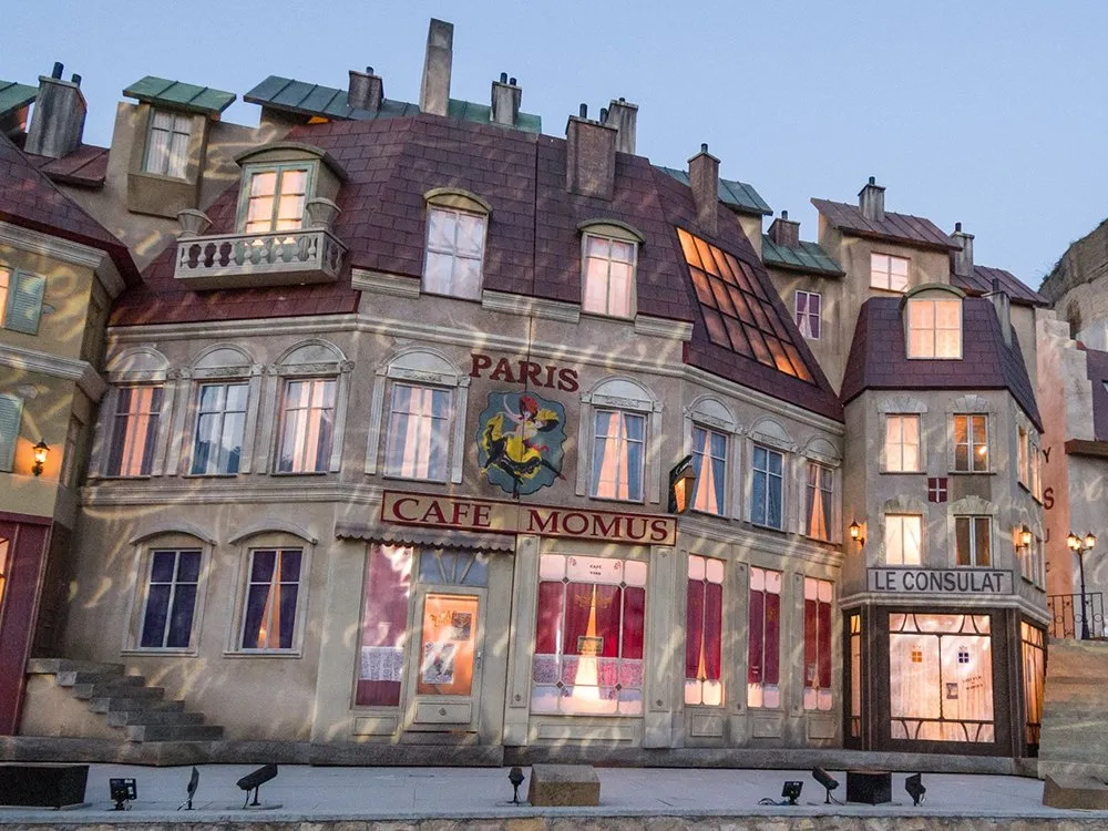 Kulisse eines pariserischen Gebäudes mit Café Momus und Le Consulat, beleuchtet mit warmem Licht an Fenstern und Laternen