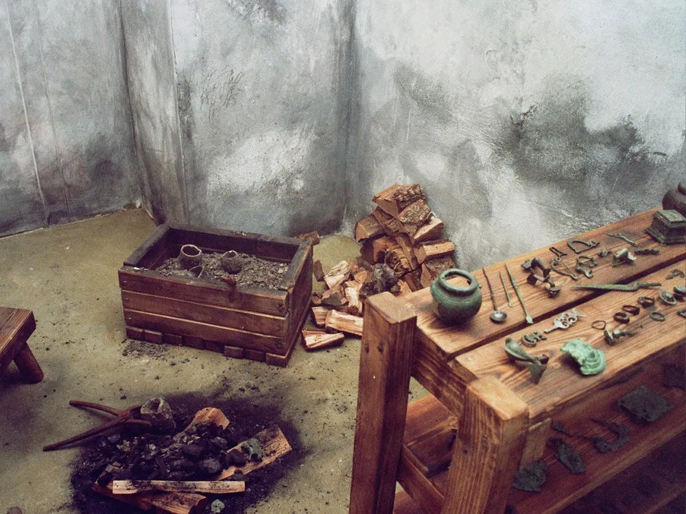 Nachbildung einer alten Werkstatt mit Feuerstelle, Holzbox, Werkzeugen und einem Tisch mit Metallgegenständen