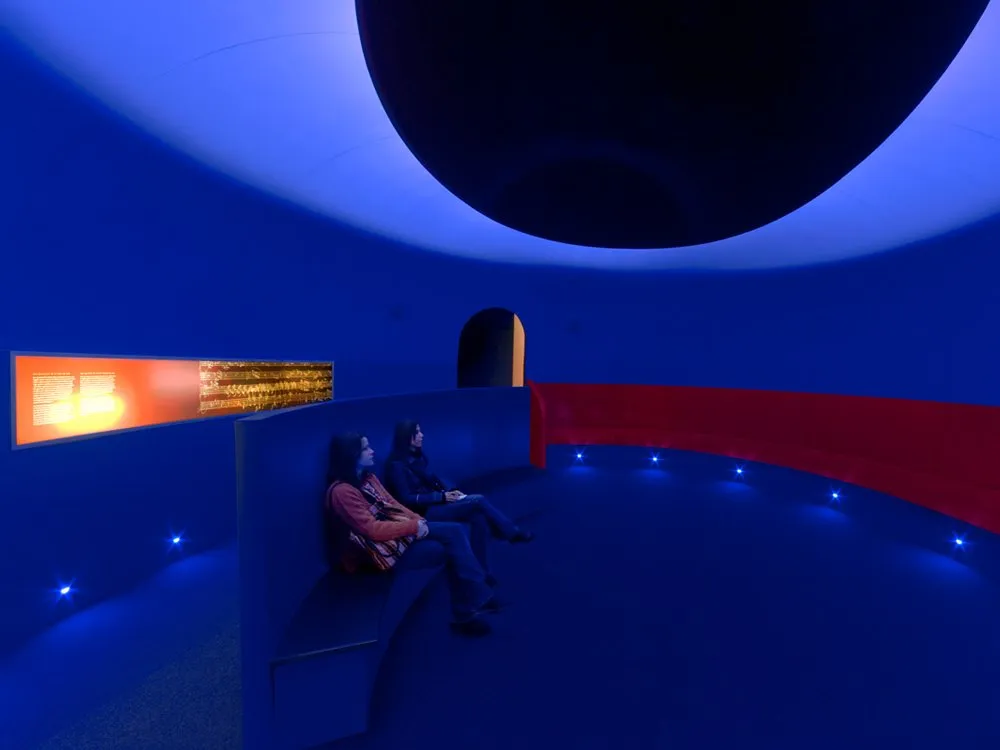Ausstellungsraum: Blauer Wand und roter Sitzbank, zwei Personen sitzen, beleuchtet mit blauen Lichtern, ein orangefarbenes Display an der Wand