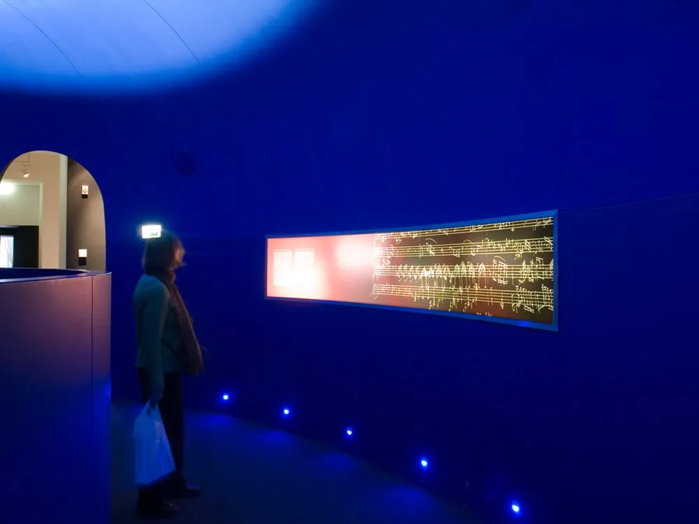 Ausstellungsraum: Blaue Wand mit orangefarbenem Display von Noten, eine Person mit Tasche steht davor, beleuchtet mit blauen Lichtern