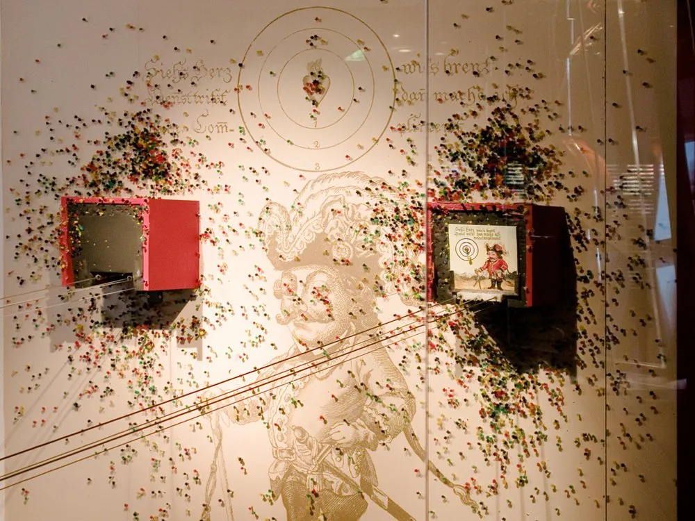usstellungsraum: Weiße Wand mit einem historischen Soldatenbild, bunte Pinnwand-Pins um die zwei roten Boxen mit Displays herum, ein Zielscheibenmotiv an der Wand