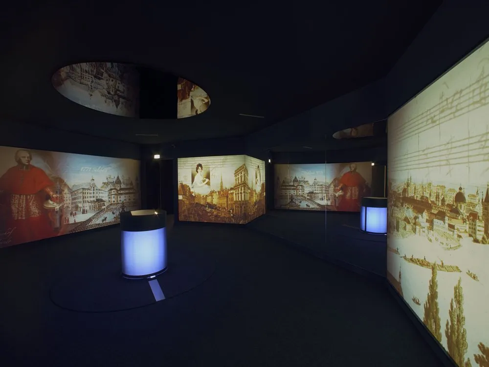 Ausstellungsraum: Dunkle Wände mit historischen Stadtbildern und Figuren, beleuchtet mit blauen Lichtern, ein zentraler runder Displaystand.