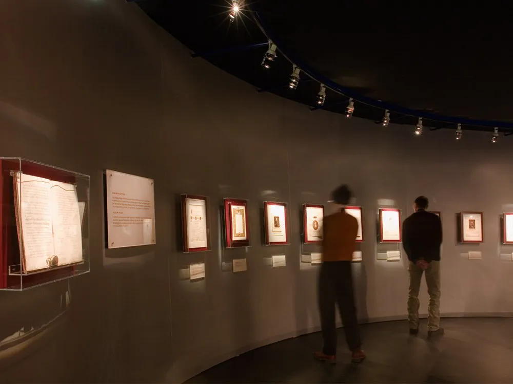 Ausstellungsraum: Graue, gebogene Wand mit Vitrinen und gerahmten Dokumenten, zwei Personen betrachten die Ausstellungsstücke
