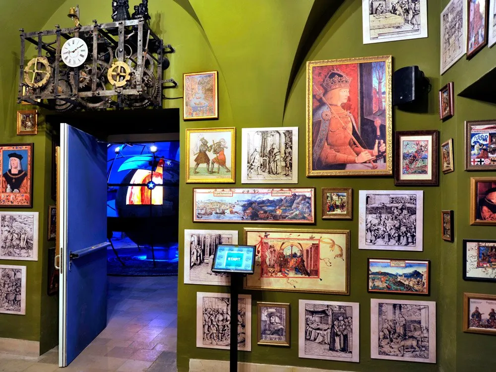 Ausstellungsraum: Grüne Wände mit zahlreichen historischen Gemälden und Zeichnungen, eine mechanische Uhr und ein interaktiver Bildschirm mit "Start"-Button.