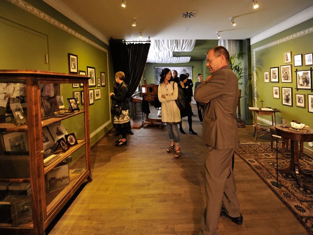 Ausstellungsraum: Grüne Wände mit alten Fotografien, eine Vitrine mit Erinnerungsstücken und Menschen, die historische Ausstellungsstücke betrachten.