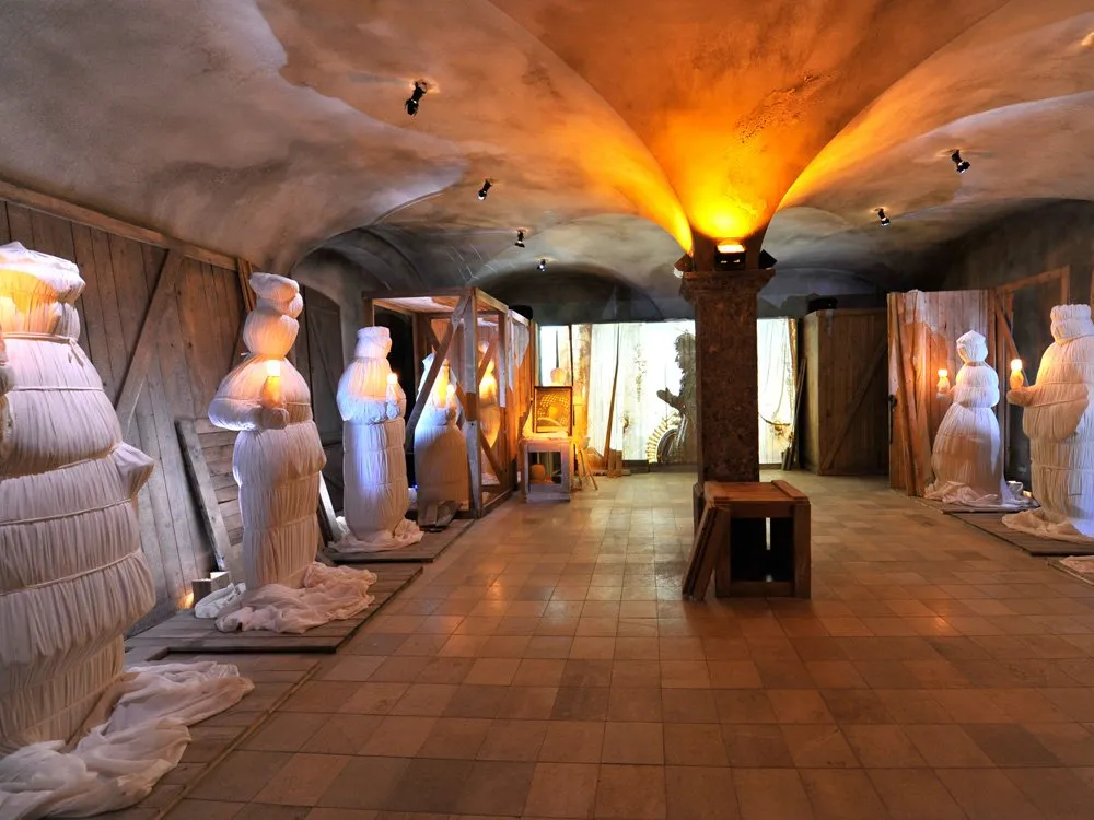Ausstellungsraum: Gewölbter Raum mit weißen, gefalteten Skulpturen, warmem Licht und einer zentralen Säule, umgeben von Holzwänden.
