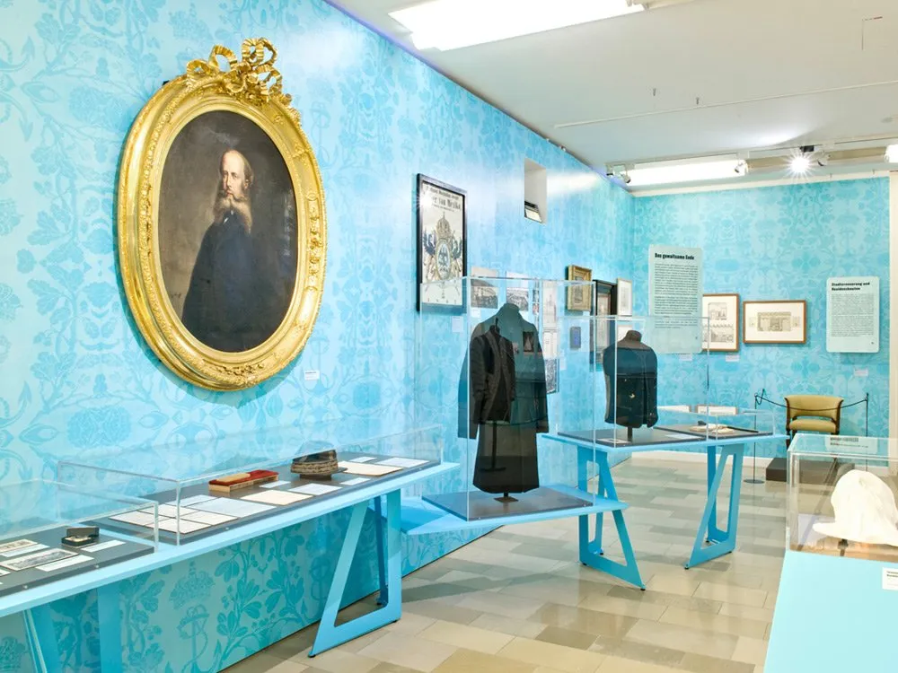 Ausstellungsraum: Blauer Raum mit floraler Tapete, goldgerahmtes Porträt, Vitrinen mit Kleidung, Dokumenten und einem Hut, an den Wänden hängen Bilder und Plakate.