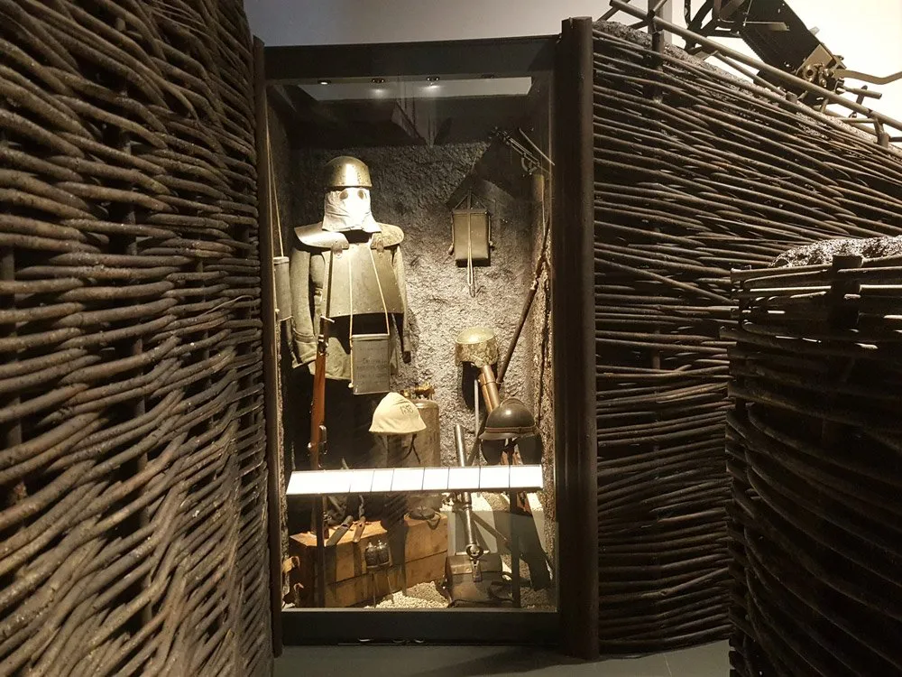 Vitrine mit historischer Militärausrüstung, darunter ein Soldat in Uniform, Helme und Kisten, umgeben von geflochtenen Wänden
