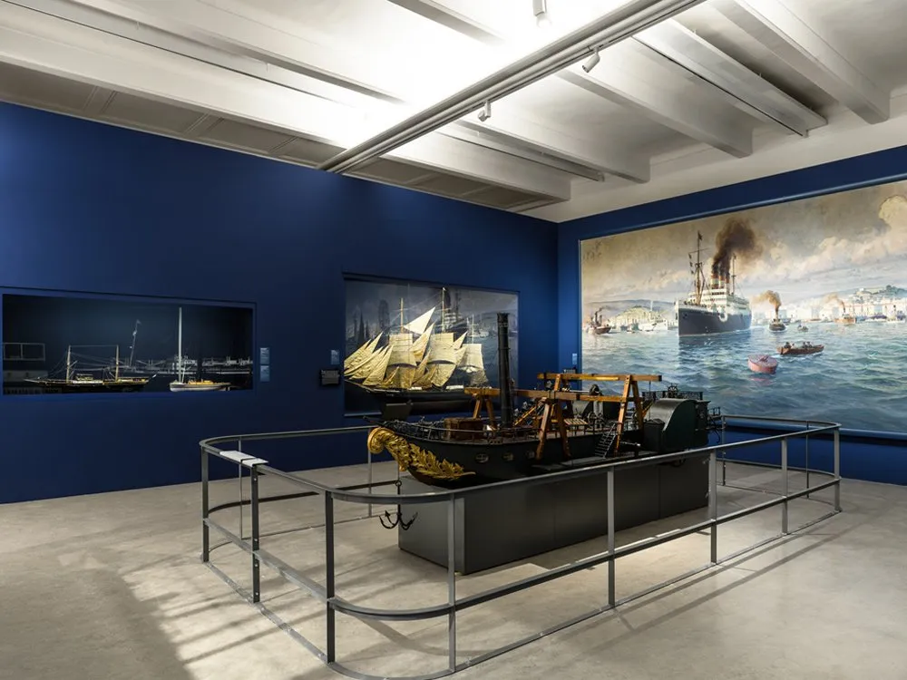 Ausstellungsraum: Blauer Raum mit einem Modellschiff und Gemälden von Schiffen an den Wänden, beleuchtet von Deckenstrahlern.