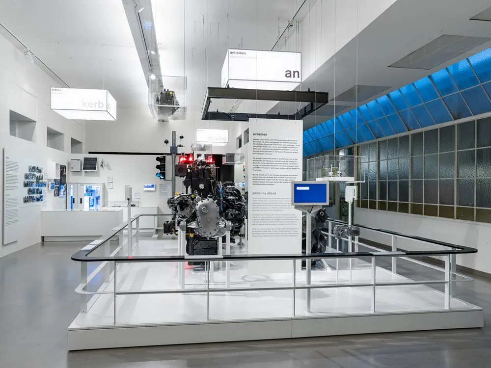 Ausstellungsraum: Weißer Raum mit einer Ausstellung von Motoren unter der Überschrift "an treiben powering", umgeben von Informationspaneelen und blauen Fenstern.