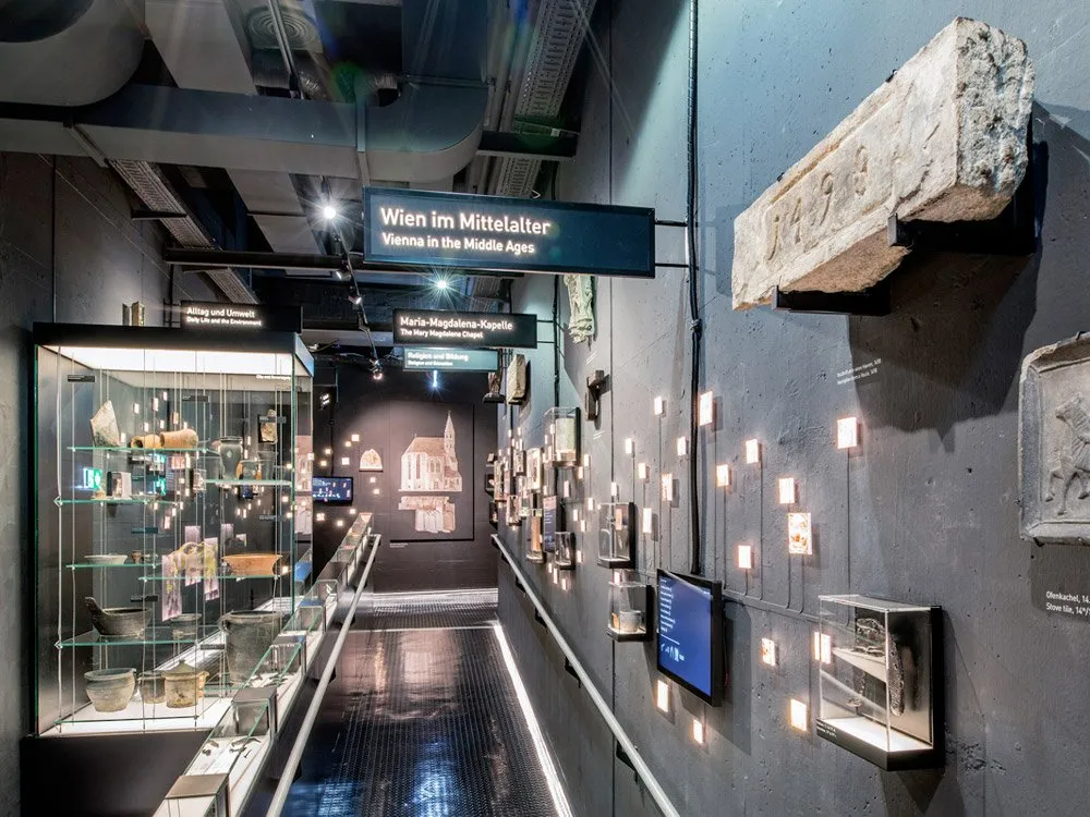 Ausstellungsraum: Dunkler Gang mit Vitrinen und Artefakten, Schildern wie "Wien im Mittelalter" und "Maria am Gestade Kapelle", sowie historischen Steinen und Bildschirmen an den Wänden