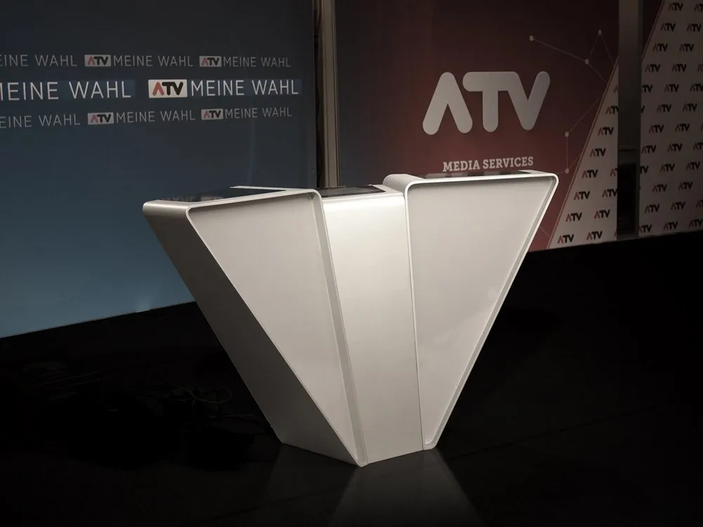 moderner Pulttisch im Fernsehstudio mit dem ATV-Logo und "Meine Wahl"-Schildern im Hintergrund