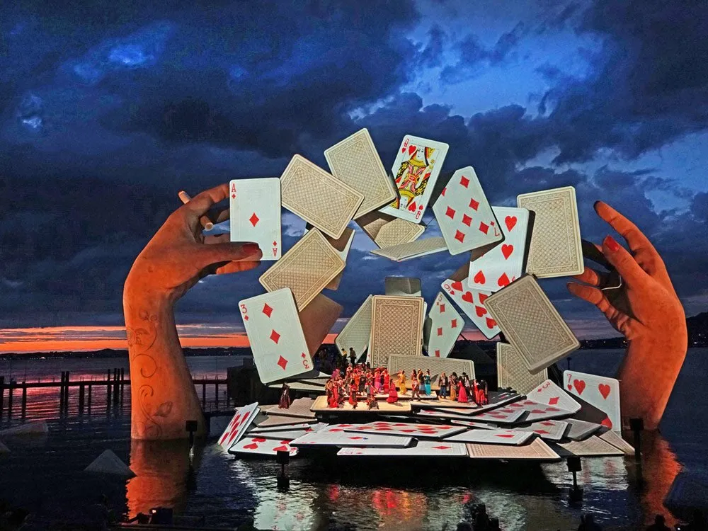Festspielbühne am See mit riesigen Spielkarten, gehalten von großen Händen, Tänzer auf der Bühne, im Hintergrund Sonnenuntergang und Wasser