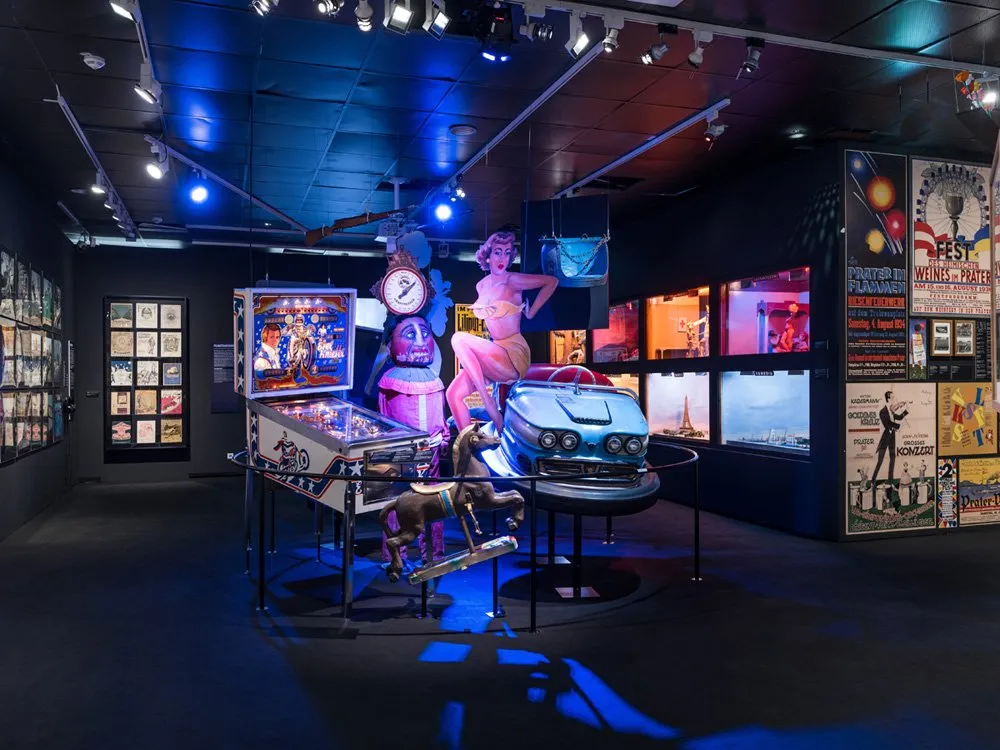 Ausstellungsraum: Flipperautomat, Karussellpferd, blaues Auto-Modell, große Figur, Poster an den Wänden, blaue Beleuchtung, dunkler Boden