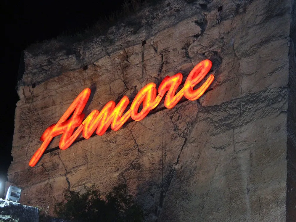 Szene: Ein Felsen bei Nacht mit einem leuchtend orangefarbenen "Amore"-Neonschild, das in den Stein eingelassen ist.
