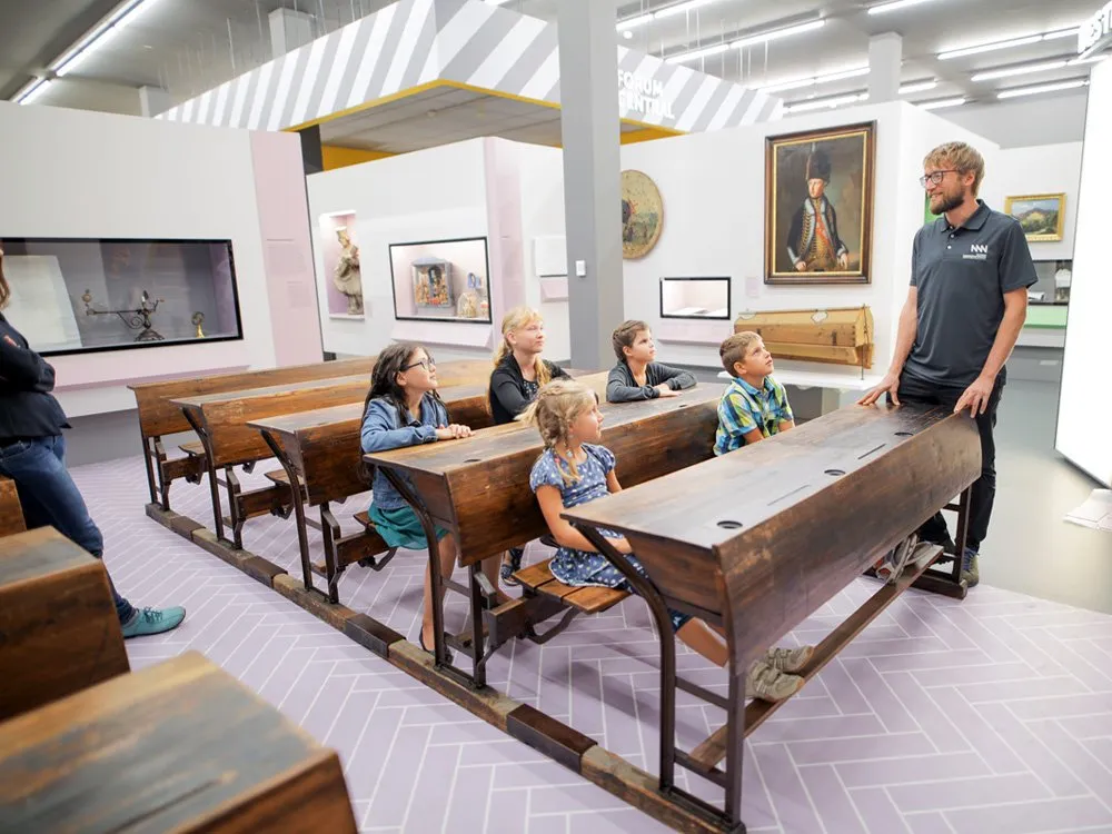 Ausstellungsraum: Kinder sitzen an alten Schulbänken, während ein Mann in Uniform sie anspricht, umgeben von Kunstwerken und Bildschirmen an den Wänden
