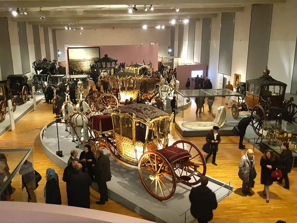 Ausstellungsraum: Prächtige historische Kutschen, darunter eine goldene mit Pferden, auf einer erhöhten Plattform, Besucher umherlaufend