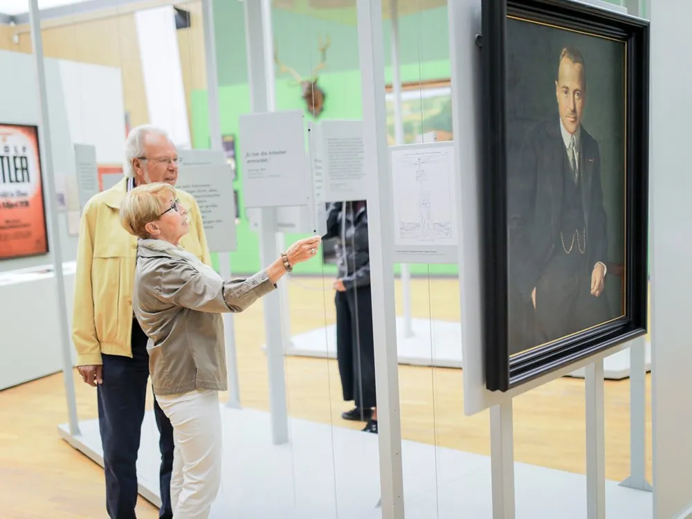 Ausstellungsraum: Zwei ältere Personen betrachten ein Porträt und Infotafeln, umgeben von grünen Wänden und einem Geweih-Dekor
