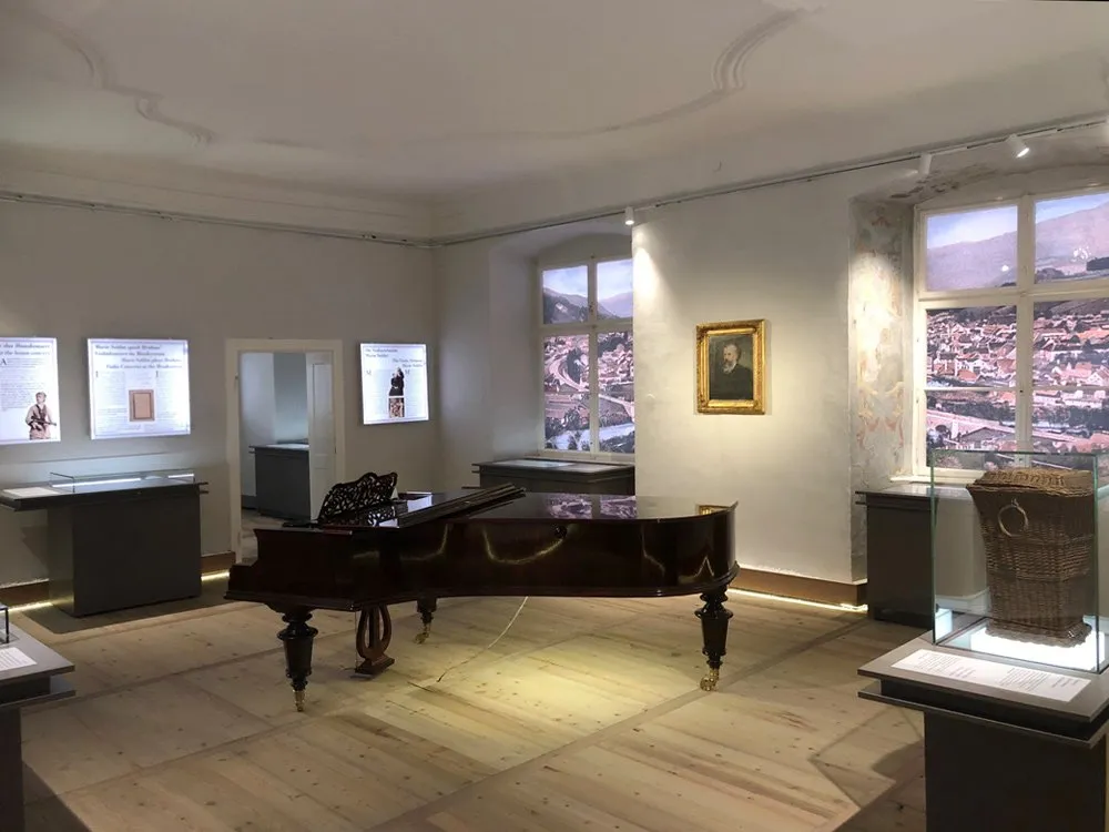 Ausstellungsraum: schwarzer Flügel steht im Zentrum, umgeben von Vitrinen mit Exponaten und Porträt an der Wand