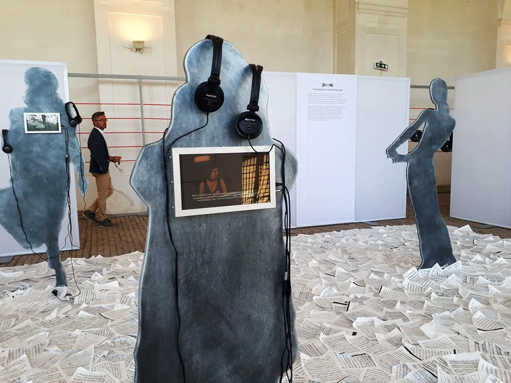 Ausstellungsraum mit abstrakten Silhouettenfiguren, die Kopfhörer tragen und kleine Bildschirme mit Fotos oder Videos enthalten.