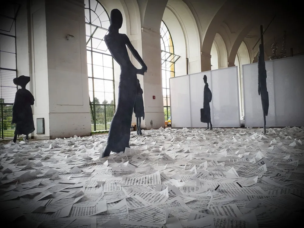 Ausstellungsraum mit abstrakten Silhouettenfiguren auf verstreuten Papieren, erleuchtet durch große Fenster