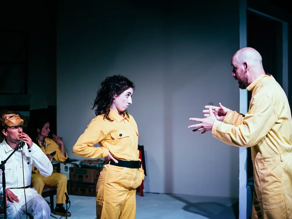 Szene: Zwei Schauspieler in gelben Overalls führen eine dramatische Konfrontation auf der Bühne, während andere Darsteller im Hintergrund sitzen und beobachten