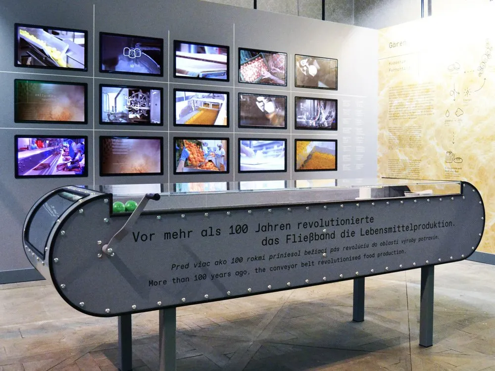 Ausstellungsraum: Ein Modell eines Förderbands mit Texten über die Revolutionierung der Lebensmittelproduktion vor über 100 Jahren, umgeben von Bildschirmen mit industriellen Szenen